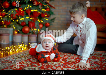 Brother with little sister dressed as Santa Claus. at the New Year tree. Family celebrates Christmas. 6 months old little boy learns to crawl on backg Stock Photo