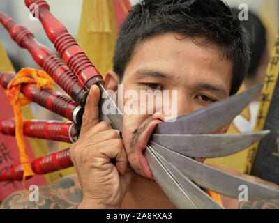 Entranced young Thai Chinese Taoist devotee (mah song) pierces his right cheek with five large knives during the Phuket Vegetarian Festival. Stock Photo
