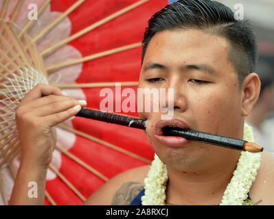Entranced young Thai Chinese Taoist devotee (mah song) pierces his right cheek with a wooden parasol handle during the Phuket Vegetarian Festival. Stock Photo