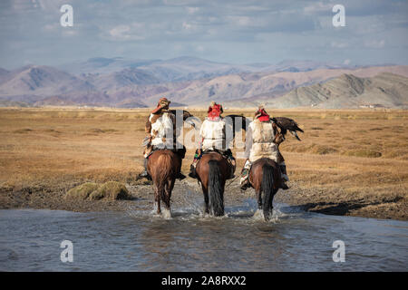 A group of traditional kazakh eagle hunters holding their golden eagles on horseback while walking through a river. Ulgii, Mongolia. Stock Photo