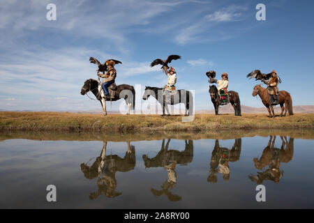 A group of traditional kazakh eagle hunters holding their golden eagles on horseback at the edge of the river with their reflections. Ulgii, Mongolia. Stock Photo