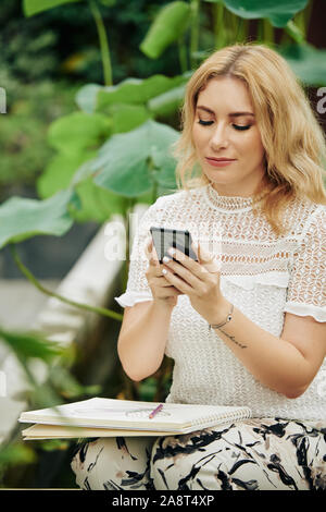 Attractive young smiling woman resting in city garden and checking messages on her smartphone Stock Photo