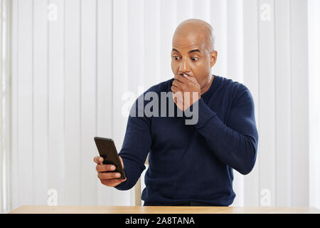 Handsome amazed fit man covering mouth with hand when reading message with shocking news on smartphone Stock Photo