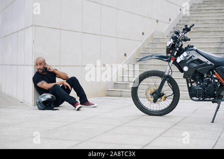 Mixed-race motorcyclist sitting on ground and looking around the building corner Stock Photo