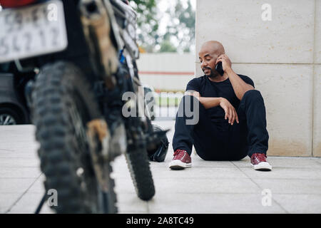 Handsome mixed-race man sitting on ground and calling mechanic as his motorcycle got broken Stock Photo