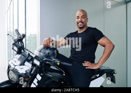 Happy confident man with white toothy smiling sitting on motorcycle with helmet in hand and looking at camera Stock Photo