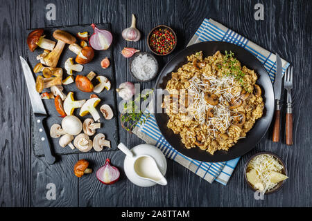 Creamy risotto with wild mushrooms, grated cheese and thyme on a black plate with ingredients on a wooden table, view from above, flatlay Stock Photo