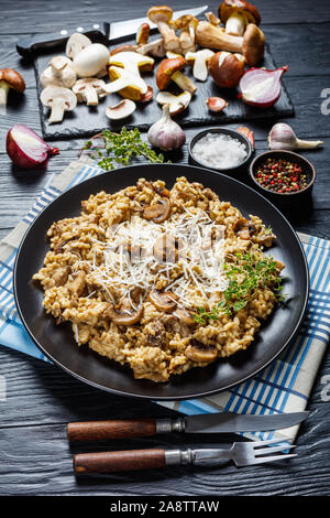 vertical view of wild mushrooms Creamy risotto with grated cheese and thyme on a black plate with ingredients on a wooden table Stock Photo