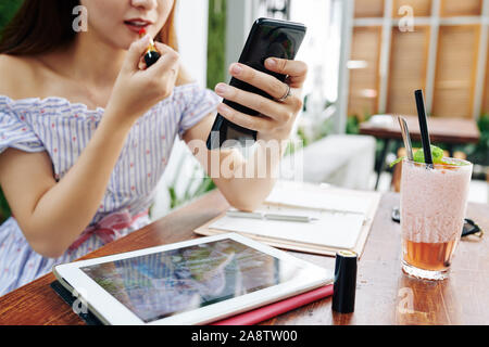 Young woman sitting at cafe table and using camera on her smartphone when applying bright red lipstick Stock Photo