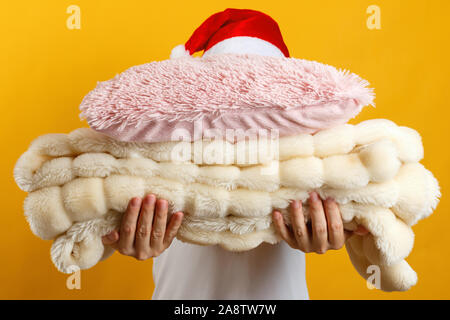 Woman in santa hat holding a stack of warm fluffy plaid and pillows on a background of a yellow wall. Stock Photo