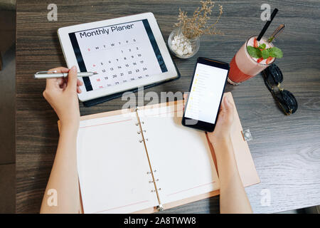 Hands of businesswoman checking monthly planner and documents in her smartphone when writing in diary Stock Photo
