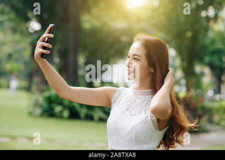 Beautiful young Vietnamese woman taking selfie on smartphone when walking in park Stock Photo