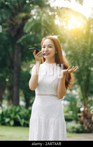 Positive young Asian woman waiting in park and recording voice message for her late friend Stock Photo