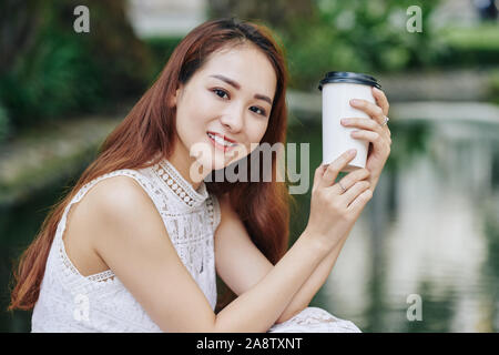 Portrait of young smiling Asian woman holding cup of take away coffee and looking at camera Stock Photo