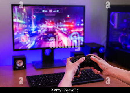 Playing racing games on computer concept. Hand holds the joystick. In the background is a gaming computer with RGB light. Stock Photo