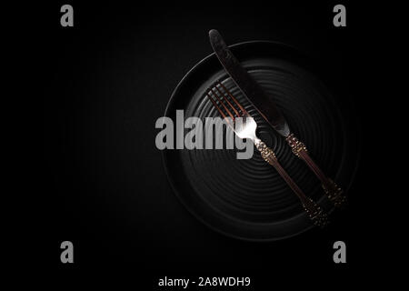 Closeup empty round black aged ceramic plate, vintage cutlery, silver fork, knife on dark background with copy space. Concept modern shooting menu, re Stock Photo