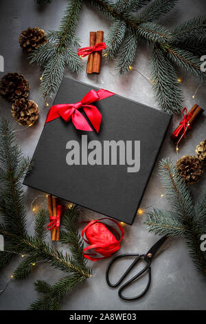 Packing Christmas gift. Festive mood flat lay with a black gift box in the middle, surrounded by the fir tree branches and red details Stock Photo