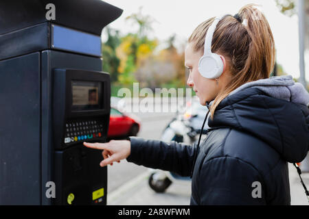 Young blonde woman paying on parking meter in the city with surprised face wearing a jacket an headphones Stock Photo