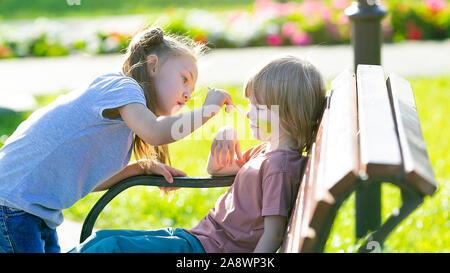 A little girl is applying aqua makeup to the face of a 5 year old boy who is sitting on a bench a park. Stock Photo