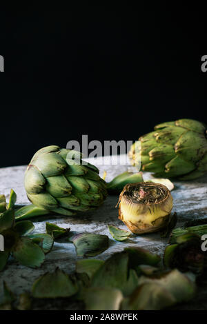 closeup of some whole raw fresh artichokes and some other cut artichokes and their leaves on a white rustic wooden table, against a black background Stock Photo