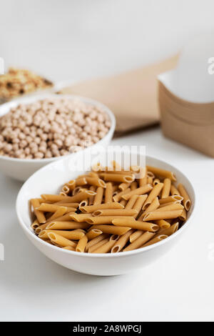 a white ceramic bowl with some penne rigate on a white table, next to a bowl with dry chickpeas a metal bowl with some walnuts and a pile of paper con Stock Photo