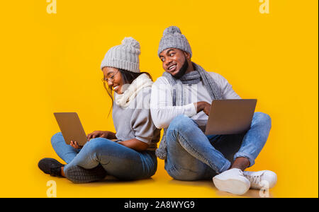Cheerful black millennial couple in winter hats browsing on laptops Stock Photo