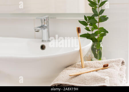 Two brown bio-degradable, compostable bamboo toothbrushes on a towel in a bathroom white interior. Green plant decor in background. Stock Photo