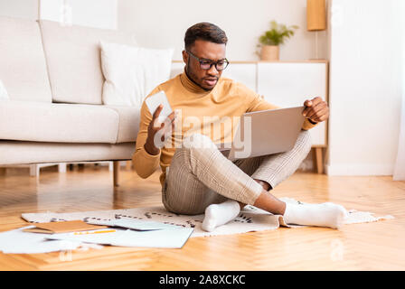 Black Man Holding Phone Working On Laptop On Floor Indoor Stock Photo