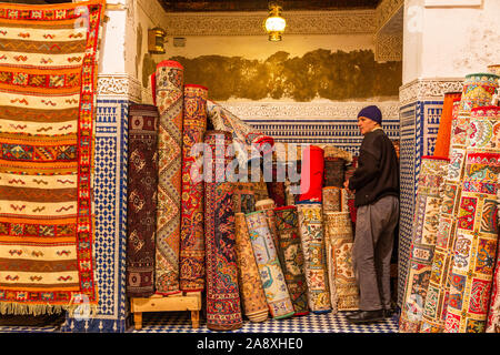 Carpet seller arranging colourful traditional rugs inside souk shop, Marrakesh Medina, Morocco, showcasing handcrafted textile heritage. Stock Photo