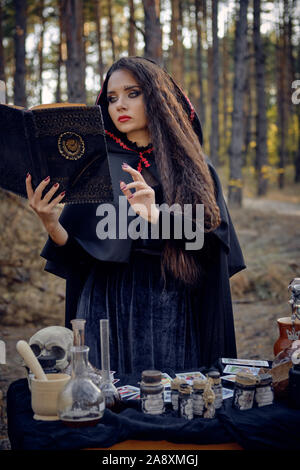 Gorgeous, wicked, long-haired sorceress in a black, long dress with cape and hood. She is holding a magic book and pronouncing a spell while posing in Stock Photo