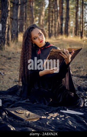Pretty, wicked, long-haired sorceress in a black, long dress with cape and hood. She is reading a book, sitting on a dark blanket whith runes and cand Stock Photo