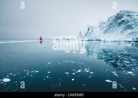 Beautiful red sailboat in the arctic next to a massive iceberg showing the scale. Cruising among floating icebergs in Disko Bay glacier during midnigh Stock Photo