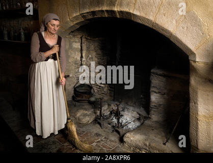 Woman dressed in renaissance maid costume using her broom to clean the fireplace in the kitchen of a property released authentic medieval castle in Fr Stock Photo