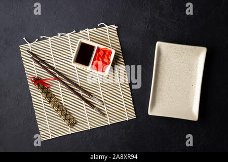 Sushi chopsticks, soy sauce, ginger, bamboo mat and empty plate on black background. Top view. Flat lay. Stock Photo