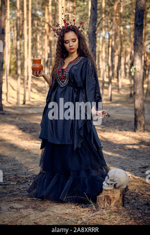 Gorgeous, wicked, long-haired sorceress in a black, long embroidered dress. There is large red crown in her brown, curly hair. She is giving some wate Stock Photo