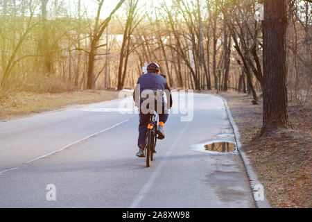 Cyclist in protective. Riding on bike in park alley on sunny day among many trees. Sunlight. Stock Photo