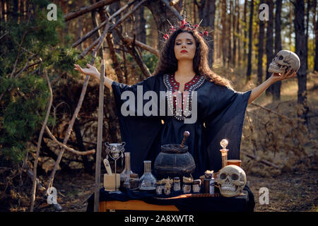 Gorgeous, wicked, long-haired sorceress in a black, long embroidered dress. There is large red crown in her brown, curly hair. She is holding a skull Stock Photo