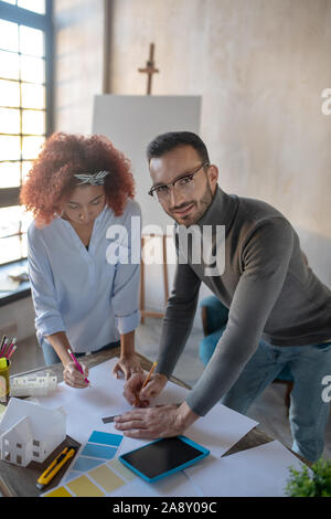 Dark-eyed interior designer making sketches with colleague Stock Photo