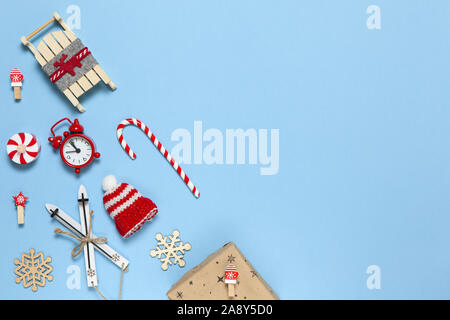 Corner creative Christmas composition. Candy cane, gift in craft paper, sled with deer, hat, alarm clock, ski, clothespins, wooden snowflakes on blue Stock Photo