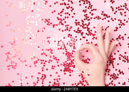 Beautiful female hand holding red foil heart with vibrant red sparkling heart-shaped confetti poured on pink background. Flat lay. Love and celebratio Stock Photo