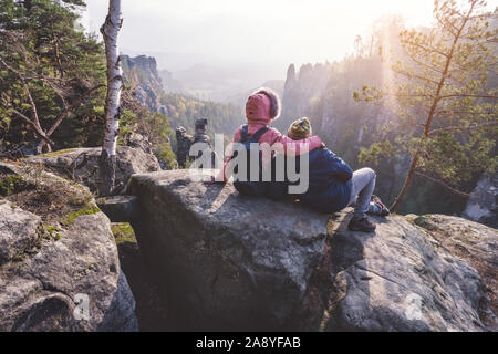Young couple in outdoor clothing with backpacks resting after hike on limestone rock enjoying back lit view of mountain ridge and forest down the Stock Photo