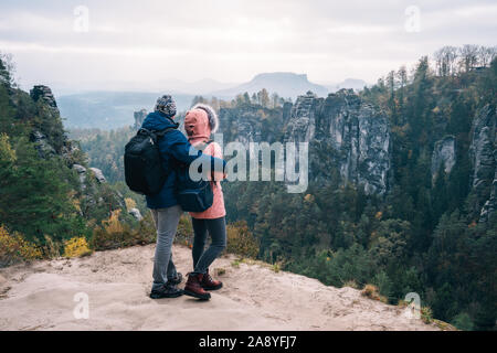 Young couple in outdoor clothing with backpacks standing on plateau enjoying view of mountain ridge and wild forest in the valley on hiking trail Stock Photo