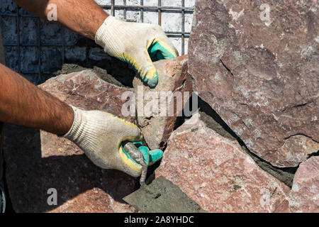 Builder - mason hands placing stones on a wall using a spatula with cement, outdoors Stock Photo
