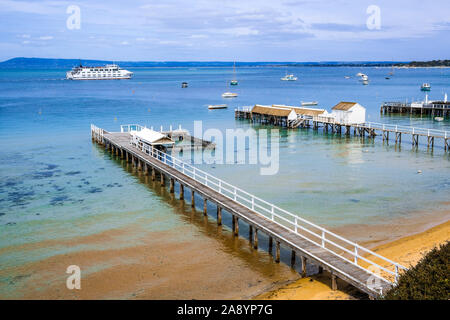 Coastal landscape - wooden piers extending into shallow bay water with passenger ferry sailing in the distance. Melbourne, Victoria, Australia Stock Photo