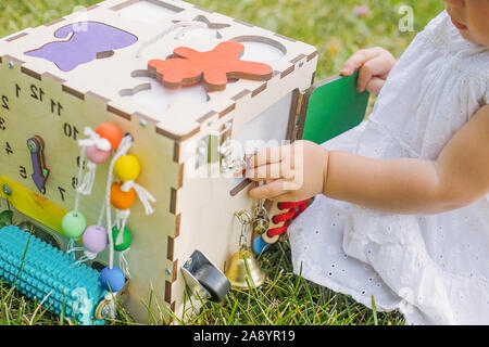 Cute little girl is playing with busiboard outdoors on green grass. Educational toy for toddlers. girl opened door to cube of board Stock Photo