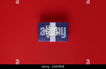 Gift box in the center of the frame on a red background. A box of blue in sparkles and with a snowflake. View from above. Minimalism. flat lay. Stock Photo