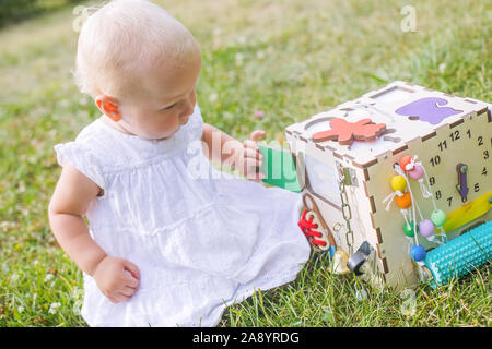 Cute little girl is playing with busiboard outdoors on green grass. Educational toy for toddlers. girl opened door to cube of board Stock Photo