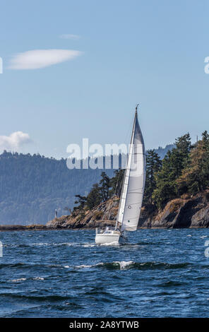 Under clear skies and amid breaking waves, a man sails a boat on a beam reach, offshore of a rocky headland on Salt Spring Island, British Columbia. Stock Photo