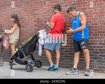 Three people standing in front of a brick wall using their phones in Harvard Square Stock Photo