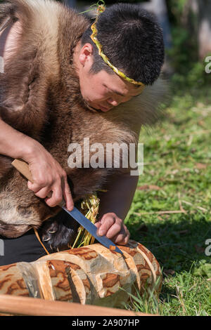 Man in traditional clothing aboriginal of Kamchatka Peninsula carved idol face with knife from birch tree during Itelmens national ritual festival Alh Stock Photo